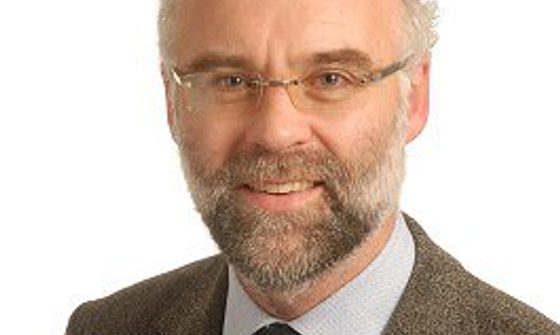 Headshot of Jon wearing a brown jacket, grey shirt and brown tie. He also wears glasses and has a beard