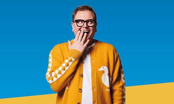 Alan Carr with a cheeky grin covering his mouth and looking towards the top right corner. He's wearing a white t-shirt with a mustard yellow cardigan that has a white diamond pattern down the sleeves and a white duck on the front