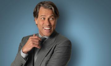 John Bishop wearing a grey suit smiling with his hand near his chin