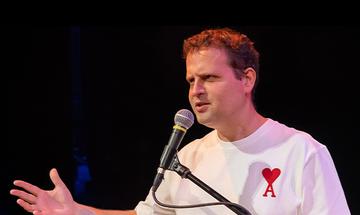 Adam Kay wearing a white t-shirt with a red heart and the letter A on, in front of a microphone