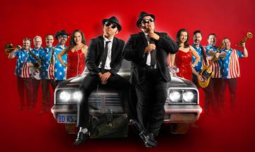 The Chicago Blues Brothers dressed in black suit and ties, sat on the front of a car. There are other musicians and singers either side, on a red background
