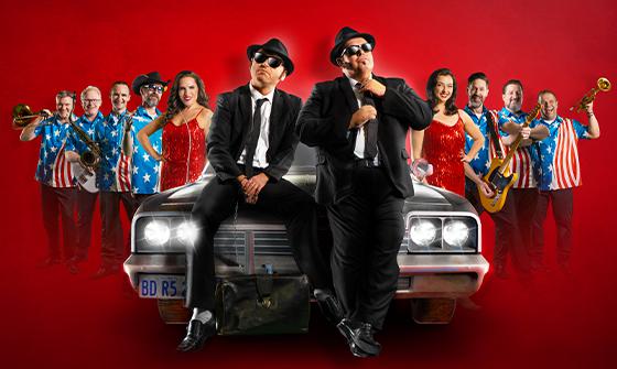 The Chicago Blues Brothers dressed in black suit and ties, sat on the front of a car. There are other musicians and singers either side, on a red background