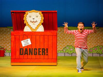 An actor from Dear Zoo performing on stage next to a pretend lion in a red box that says 'DANGER!'