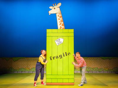 Two actors from Dear Zoo performing on stage with a pretend giraffe behind a green box that says 'fragile'