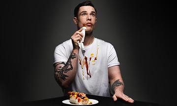 Ed Gamble sat at a table with a hotdog in front of him on a plate, with red and yellow sauces around his mouth and down the front of his white t-shirt. He has one hand on the table and the other wiping the corner of his mouth, looking up and away from the camera, on a dark background