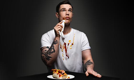Ed Gamble sat at a table with a hotdog in front of him on a plate, with red and yellow sauces around his mouth and down the front of his white t-shirt. He has one hand on the table and the other wiping the corner of his mouth, looking up and away from the camera, on a dark background