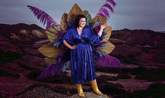 Kiri Pritchard-McLean standing on and in front of rocks wearing a blue sequined dress, gold boots, and colour peacock wings