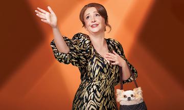 Lucy Porter waving one arm in the air and the other holding a bag that contains a dog, stood in front an orange-lit background