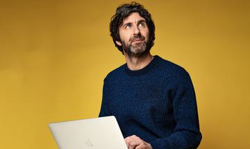 Mark Watson looking up to the sky whilst on a computer, on a yellow-gold background