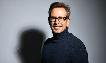 Nick Heyward smiling at the camera on a plain background, wearing a dark blue roll-neck jumper and glasses