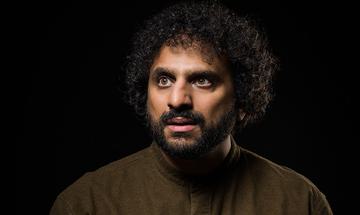 Nish Kumar with a serious face looking into the distance, on a plain black background