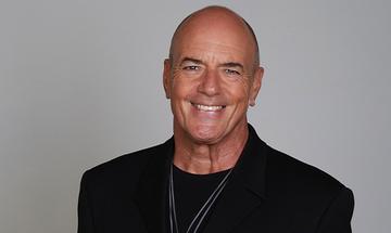 Peter Cox wearing a black jacket and smiling at the camera on a plain grey background