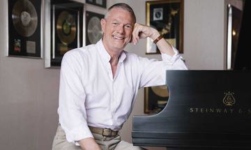 Richard Carpenter leaning on a piano, smiling at the camera