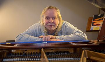 Rick Wakeman sat with his arms resting on a piano, smiling at the camera