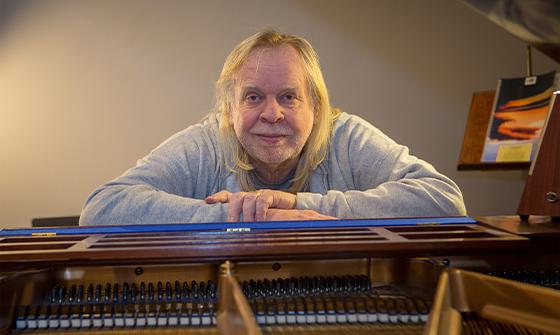 Rick Wakeman sat with his arms resting on a piano, smiling at the camera