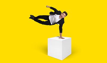 Russell Kane holding his weight on one hand on a white cubed box, with his body horizontally in the air and his mouth open