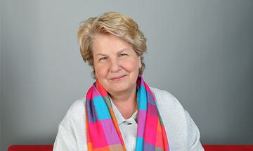 Sandi Toksvig smiling softly at the camera in a white shirt with a bright multi-coloured scarf around her. She's sat in a white room room on a red sofa