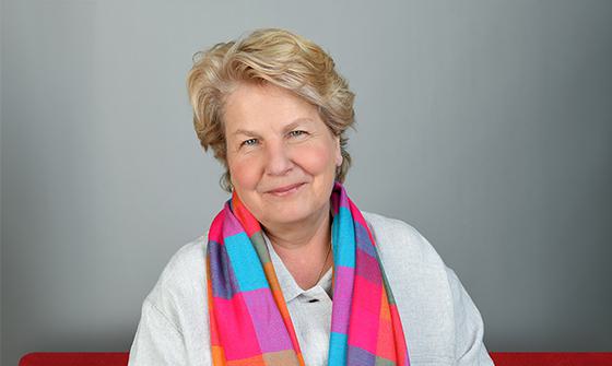 Sandi Toksvig smiling softly at the camera in a white shirt with a bright multi-coloured scarf around her. She's sat in a white room room on a red sofa