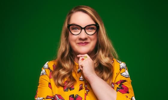 Sarah Millican wearing a yellow top smiling at the camera with her hand on her chin