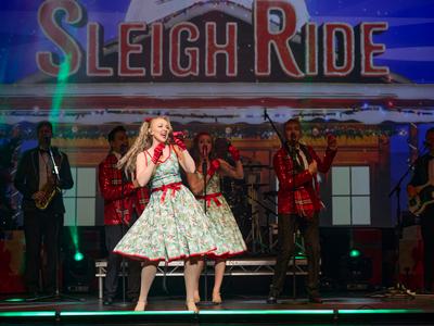 A singer in a festive dress with holly on, wearing red gloves and singing into a microphone with the words "Sleigh Ride" in the background