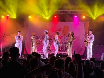 Band performing on stage, members in white outfits with gold accents under vibrant pink, yellow, and purple lights