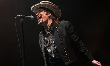 Adam Ant singing into a microphone. He is wearing a hat and a black jacket on a dark background