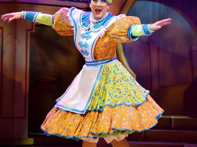 the Dame on stage with hands out to the side. She wears a bright orange, green and blue dress with a mini washing machine on her head