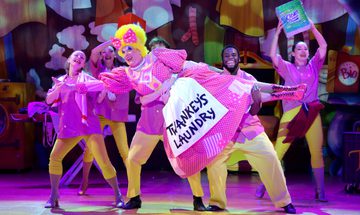 The Dame wears a bright pink frock that reads 'Twankey's Laundry'. The Dame is being held up by a few of the ensemble