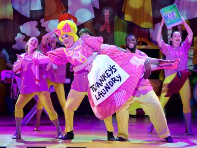 The Dame wears a bright pink frock that reads 'Twankey's Laundry'. The Dame is being held up by a few of the ensemble