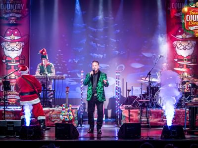 Festive music stage featuring performers in Christmas costumes, including Santa and an elf. The main singer is in a green jacket