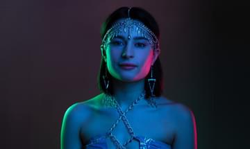 A young woman in a chain head dress with metal feathers hanging down, large triangle earrings, and chain and leather top. She is stood in front of a dark background with a blue light on her and green and red on either side