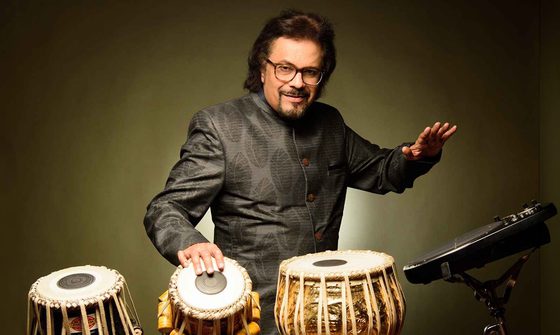 Bickram Ghosh in a black shirt with a leaf pattern smiling at the camera while playing the tabla