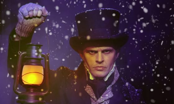 An actor dressed as Scrooge on a snowy night holding a lantern in his right hand with a stern expression on his face