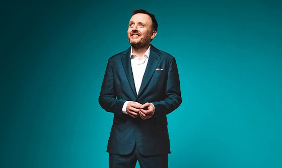 Chris McCausland standing with his hands together, wearing a dark blue suit on a blue background, smiling with his head facing slightly off camera