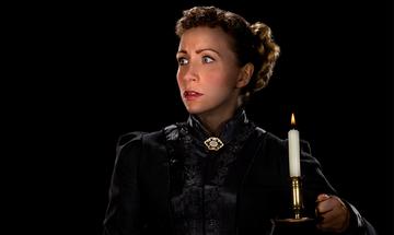 A woman with her hair tied back in a bun looking fearful in a black high necked Victorian style dress with a small gold and black brooch on the neck. She is holding a lit candle in the dark room
