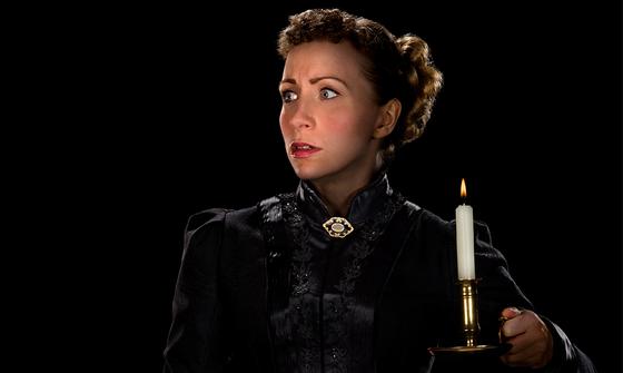 A woman with her hair tied back in a bun looking fearful in a black high necked Victorian style dress with a small gold and black brooch on the neck. She is holding a lit candle in the dark room