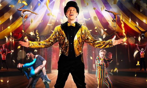 Max Fox in a black suit and top hat with gold blazer and bow tie with his arms open looking up at gold confetti. There are other performers surrounding him including acrobats, dancers, and a man holding a banana like a gun