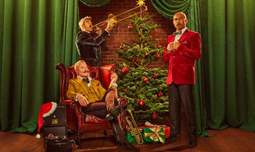 Three men on a stage with large green curtains behind them. One man with a moustache, quiffed hair, and large eyebrows is at the back playing a trombone in a black velvet suit next to a Christmas tree with a gold star at the top, red baubles, and wrapped presents at the base, alongside a trumpet. The mans eyes are very wide. A bald man with a moustache is stood on the opposite side of the tree in a red velvet suit with a and brown bow tie, he is holding his lapels looking suave. The last man is sat in a red leather armchair in front of the one with the trombone, he short blonde hair and a large fake looking black moustache. He is wearing a yellow velvet suit and matching waistcoat with a brown checked cravat, he has is legs crossed with a pipe in his hand. Next to him on the floor is a portable gramophone with a Santa hat hanging off the corner