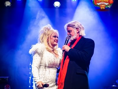 Two performers singing on stage (Dolly and Kenny tribute), illuminated by bright blue lights. The woman wears a white, feathered outfit, and the man has a dark coat and red scarf
