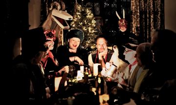 A festive photo in a house with lots of people around a dining table, being lit by a light on the table and the Christmas tree that sits behind. At the head of the table is Eliza Carthy in a black long sleeve top with a black fur hat, and Jon Boden who is in a white t-shirt an brown waistcoat with a thin red scarf. Eliza is laughing and Jon is smiling. Other people at the table is three older men shrouded in darkness in the foreground, another older man in a similar outfit to Jon, but with the addition of a top hat and glasses, he is drinking from a tankard with a goose sat between him and Jon. There is also a man dressed in all red with a demonic mask on his forehead next to Eliza, a large goat like puppet, similar to a pantomime horse behind her, and a man in all black with a homemade red mask with beads strung round it and two large sticks coming out behind Jon.