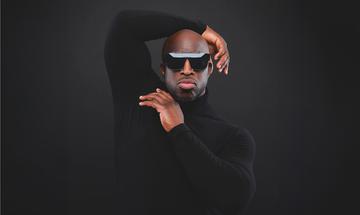 An image of Emmanuel Sonubi in a long sleeve black top with large sunglasses posing on a black background with an orange line at the bottom. He has his left arm bent over his head and his right hand under his chin, it looks like he's vouging.