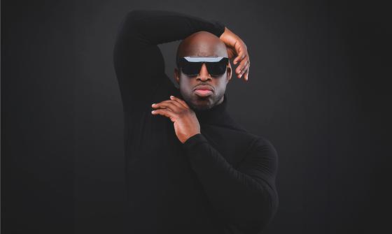 An image of Emmanuel Sonubi in a long sleeve black top with large sunglasses posing on a black background with an orange line at the bottom. He has his left arm bent over his head and his right hand under his chin, it looks like he's vouging.