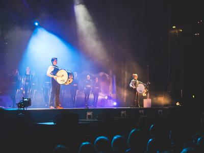 Two men standing either side of a stage each playing a drum, with multiple female dancers in all-black outfits in the background