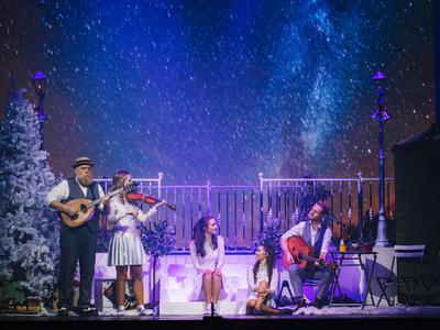 A festive stage scene with musicians playing a ukulele and violin, surrounded by a snowy tree and colorful lights against a starry backdrop