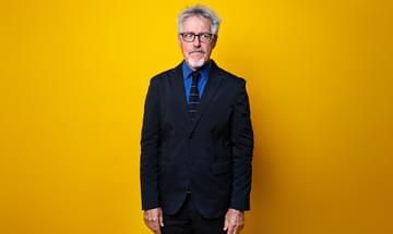 Griff Rhys Jones, a man with grey hair and glasses, standing in front of a vibrant yellow background. He is wearing a black suit, a blue shirt, and a black tie with blue stripes, looking away from the camera