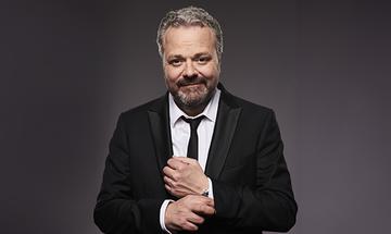 Hal Cruttenden with a beard and grey hair, dressed in a black suit and tie, smiling while adjusting his shirt cuff. The background is a neutral grey