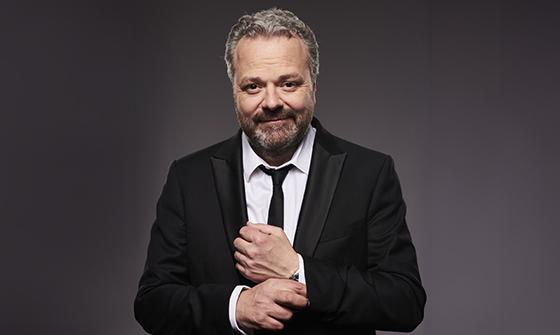 Hal Cruttenden with a beard and grey hair, dressed in a black suit and tie, smiling while adjusting his shirt cuff. The background is a neutral grey
