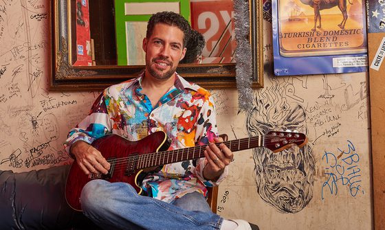 Hamilton Loomis sat in front of a wall covered in pen graffiti with a mirror and poster behind him. He is in a light multi-coloured button up shirt with blue jeans and white shoes. He has one leg crossed over the other holding his dark red acoustic guitar in his lap