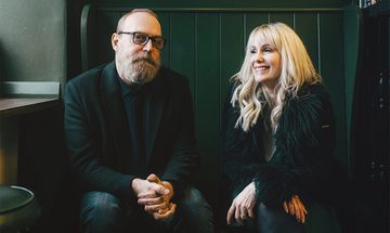 Boo Hewerdine and Heidi Talbot sat down in a dark green room. They're both in dark clothes. Boo is looking at the camera holding his hands in his lap, Heidi is looking off to the left smiling