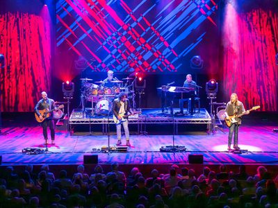 Three men playing guitars stand in a line across the front of the stage. Behind them are other musicians and a projection of red and blue lines with diagonally cross over each other.